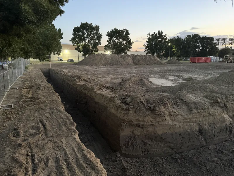 Footing trench cut square through the compacted pad, spoil windrowed alongside for backfill