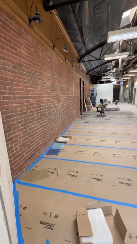 Commercial interior mid build-out with exposed brick, temporary lighting and taped floor protection covering the finished slab