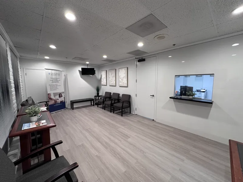 Dermatology clinic reception area with a glazed reception window, wall-mounted screen and framed artwork above patient seating