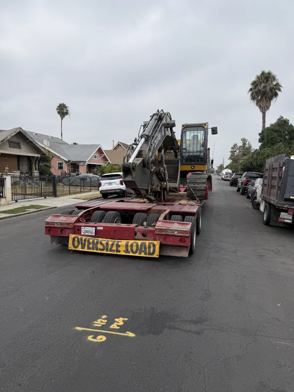 Excavator loaded on a lowboy trailer with an oversize load banner, staged in the street outside a residential demolition job