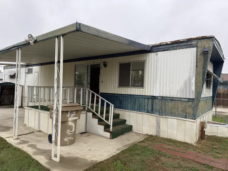 Single-wide mobile home photographed from the porch side before removal, with the original awning and steps
