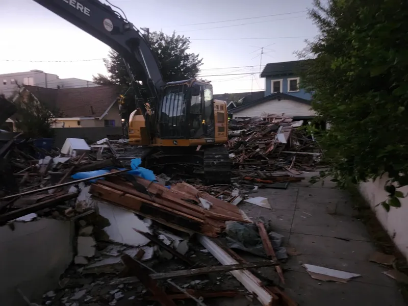 Tracked excavator standing on a demolished house working the debris pile down into the driveway at dusk