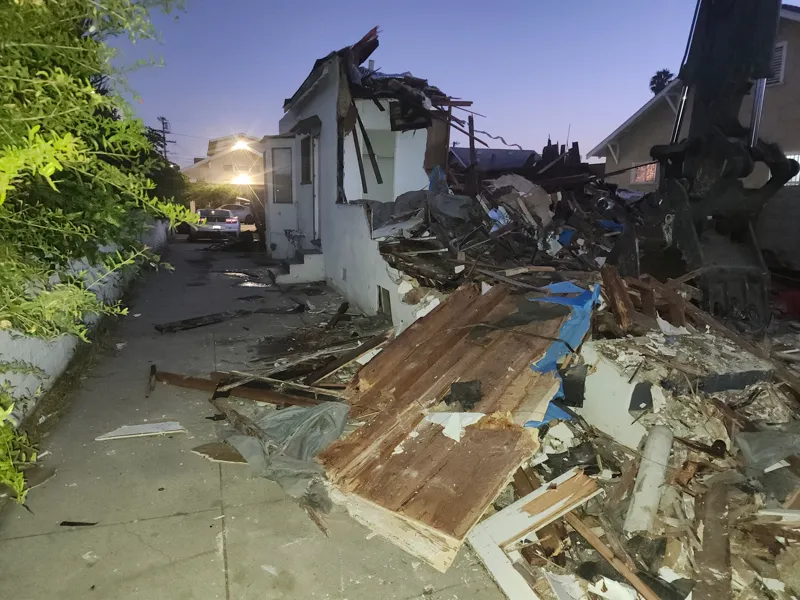 Partially demolished house at dusk with the rear half down, the excavator arm in frame and the side yard kept clear for access
