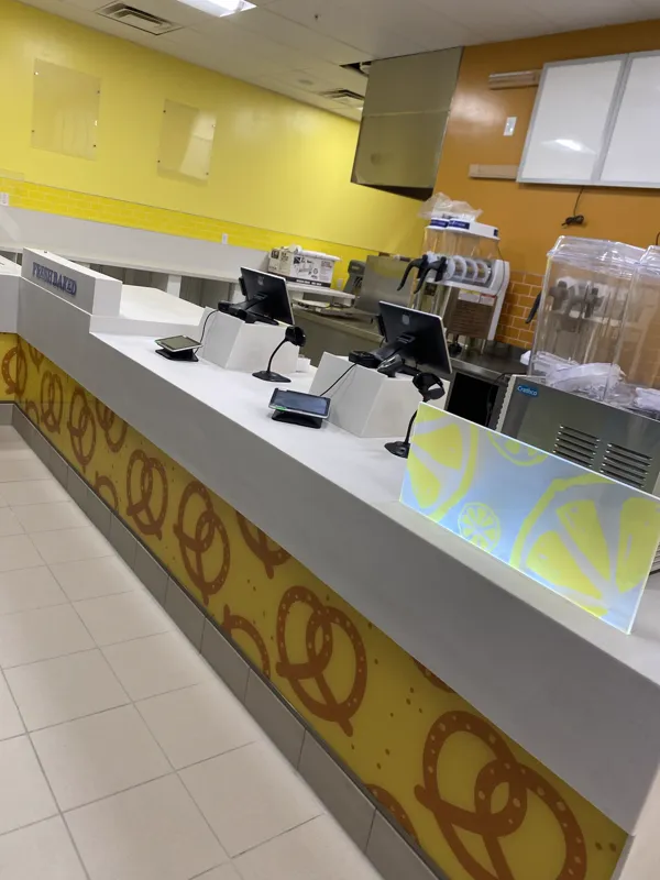 Completed food-court service counter with a quartz top, printed pretzel graphic panel, point-of-sale terminals and a frozen drink machine